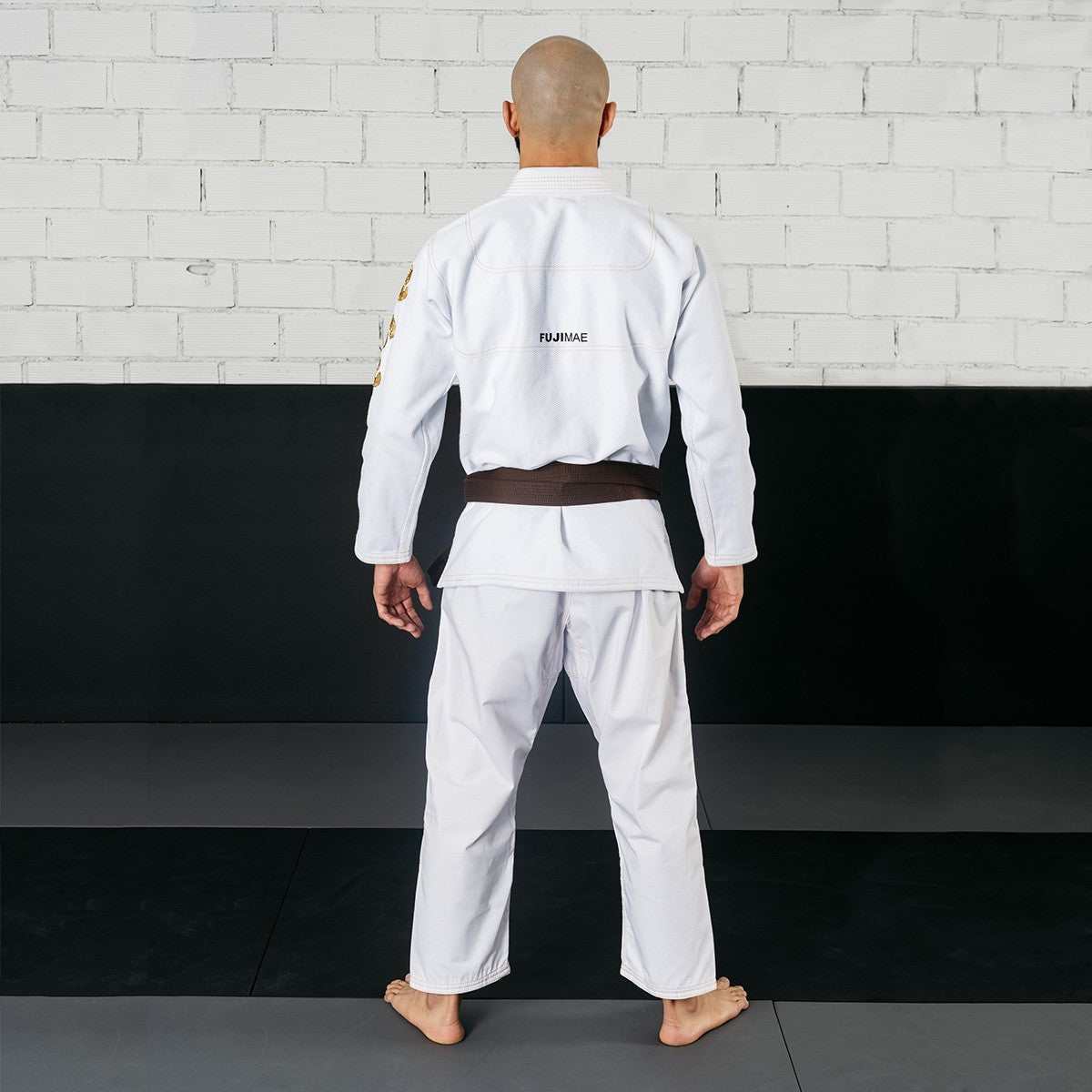 Back view of a person wearing the white Shaka 21 BJJ Gi QS with a brown belt, standing barefoot on a mat against a black and white brick wall background