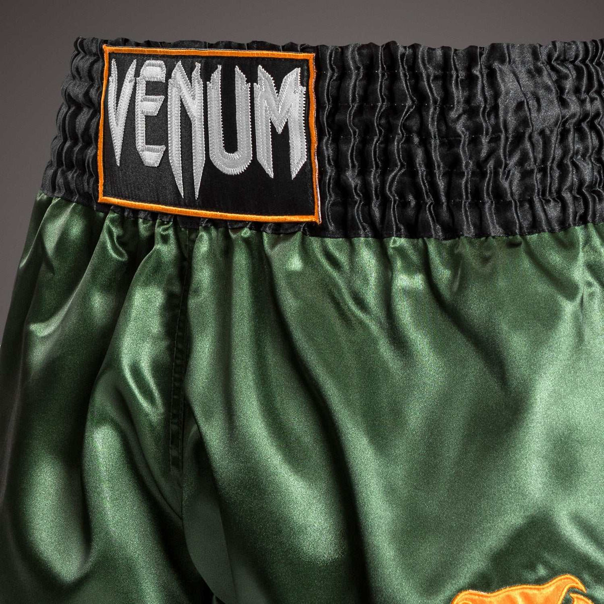 Short de Muay Thai Venum vert en polyester satiné, avec large ceinture noire et écusson logo blanc bordé d’orange.